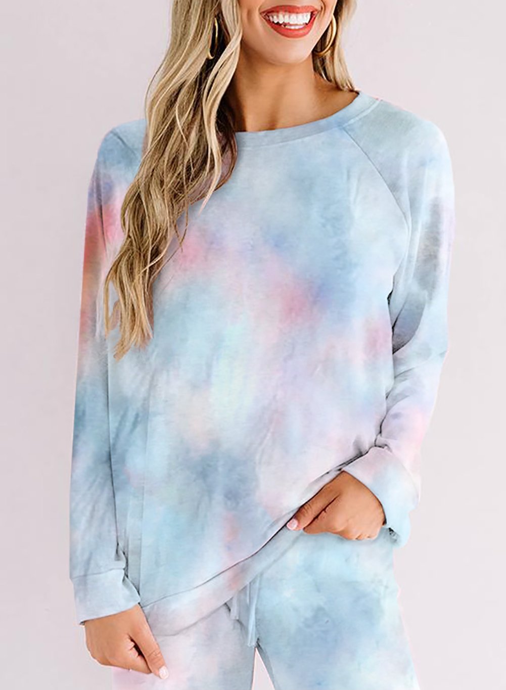 Close-up of a woman wearing a soft pastel tie-dye loungewear set in blue, pink, and lilac tones. The outfit includes a long-sleeve top and drawstring pants. She has one hand in her pocket and is smiling, with loose wavy blonde hair and hoop earrings.