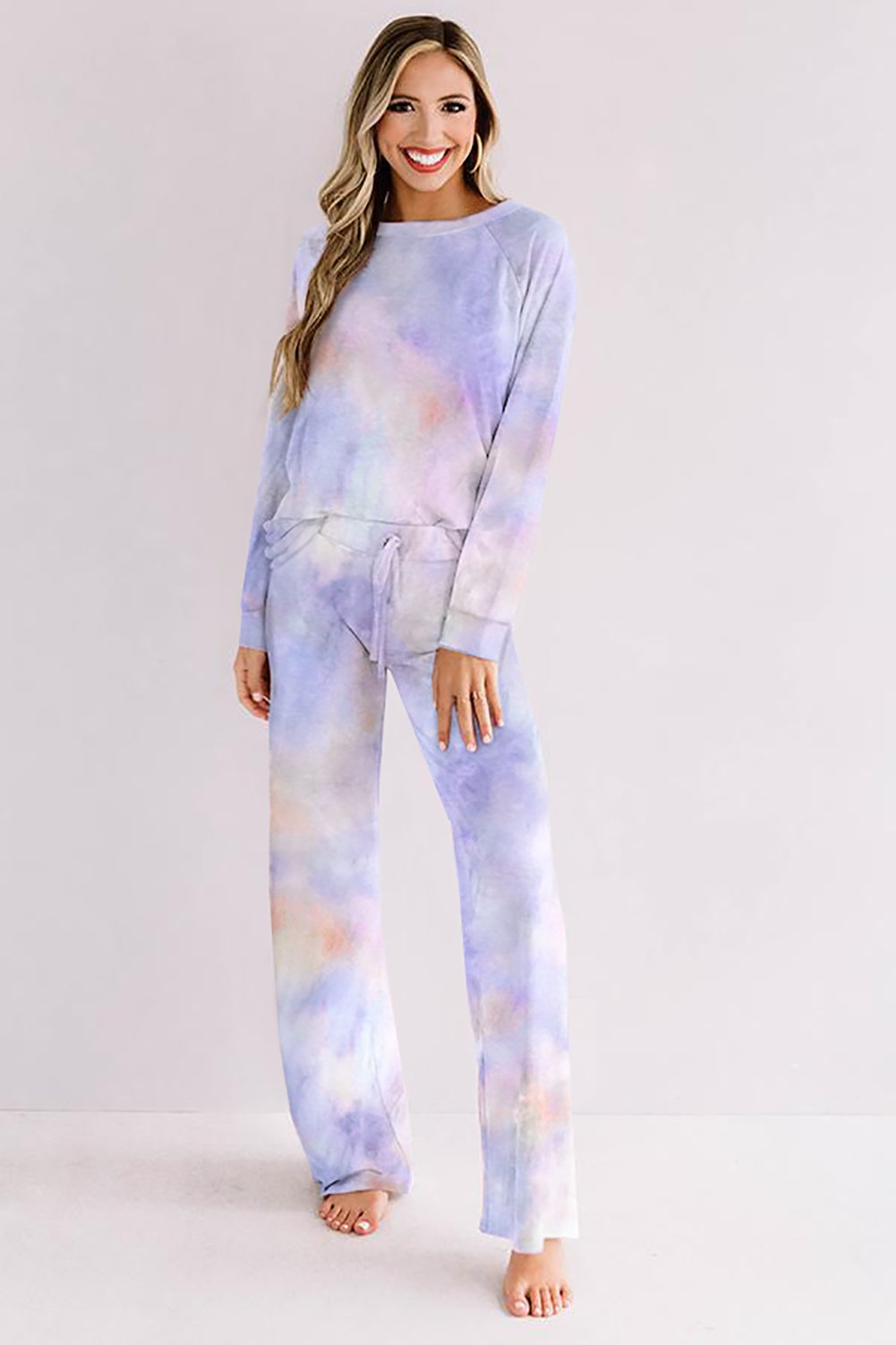 Woman smiling and posing in a relaxed-fit lounge set with a soft pastel tie-dye pattern in lavender, blush, and peach tones. The set includes a long-sleeve top and matching drawstring pants, styled barefoot against a light, minimal background.