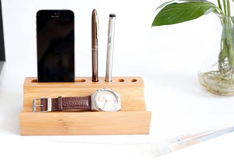 Minimalist bamboo desk organizer with slots holding a smartphone, two pens, and a wristwatch — perfect eco-friendly accessory for workspace organization.