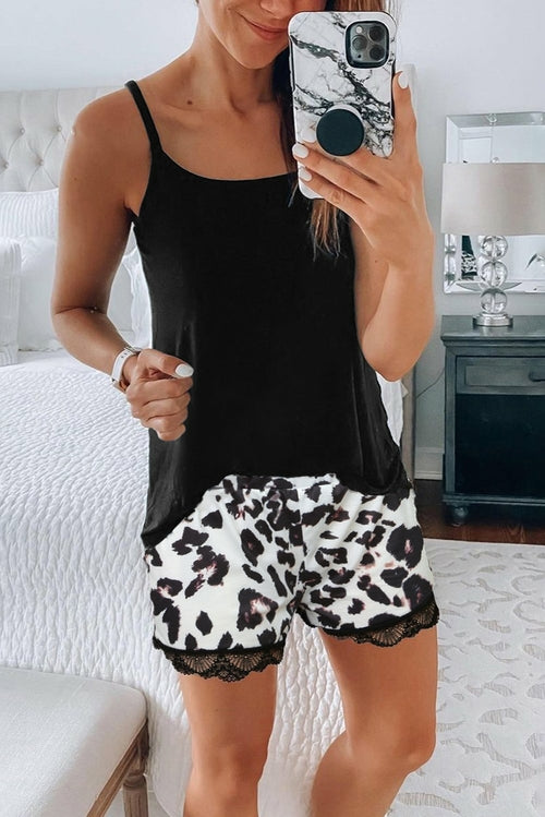 Smiling woman taking a mirror selfie in a bedroom, wearing a black camisole and white leopard-print shorts with black lace trim, standing barefoot on a white bedspread.