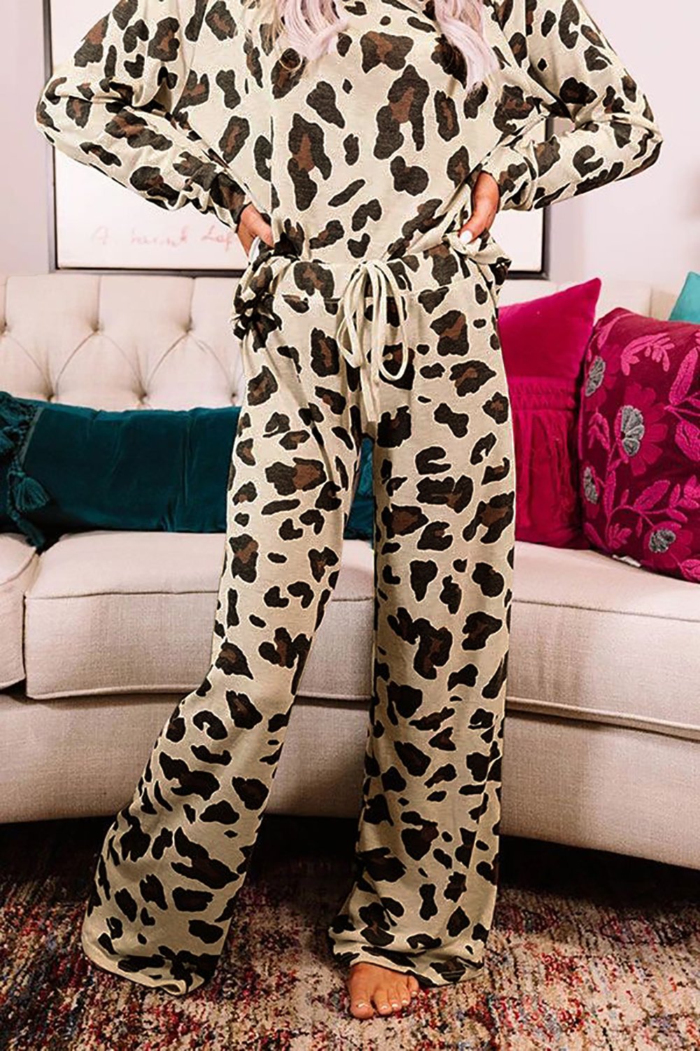 Woman wearing a beige and brown leopard print loungewear set with a long-sleeve top and wide-leg drawstring pants. She is standing barefoot on a rug in front of a cream-colored sofa with colorful cushions.