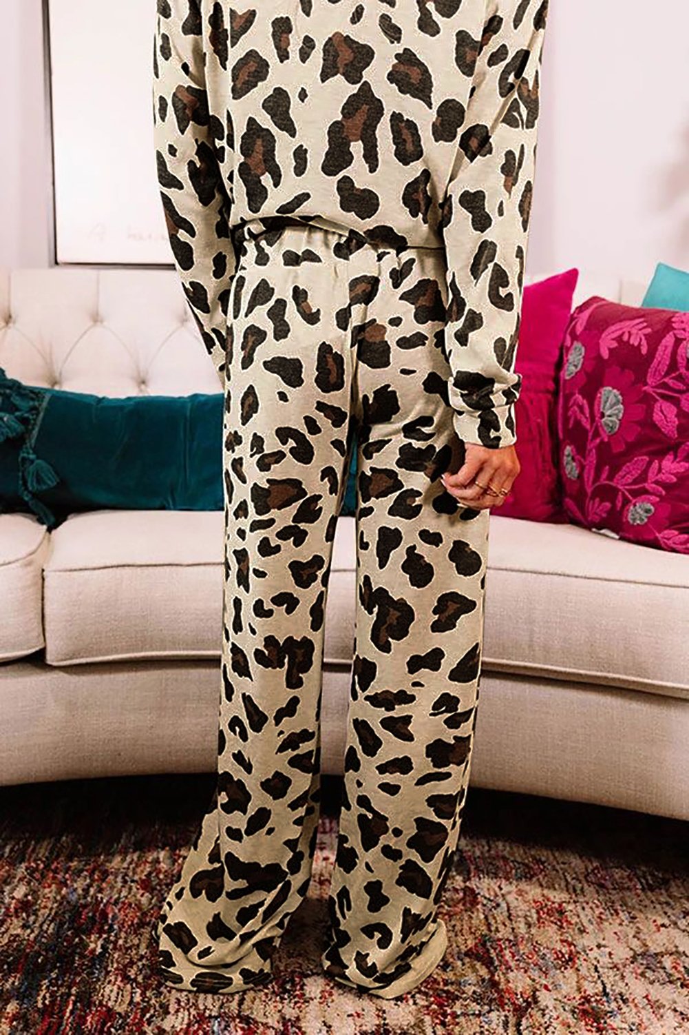 Back view of a woman wearing a beige loungewear set with an all-over dark brown and black leopard print, featuring a long-sleeve top and wide-leg pants. She is standing barefoot on a patterned rug in front of a beige sofa with colorful cushions.