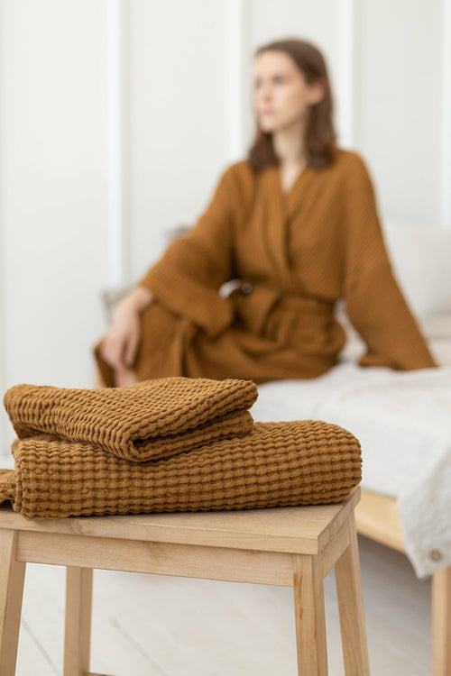 Folded amber waffle-textured robe and towel set placed on a wooden stool, with a woman in a matching robe seated in soft focus in the background.