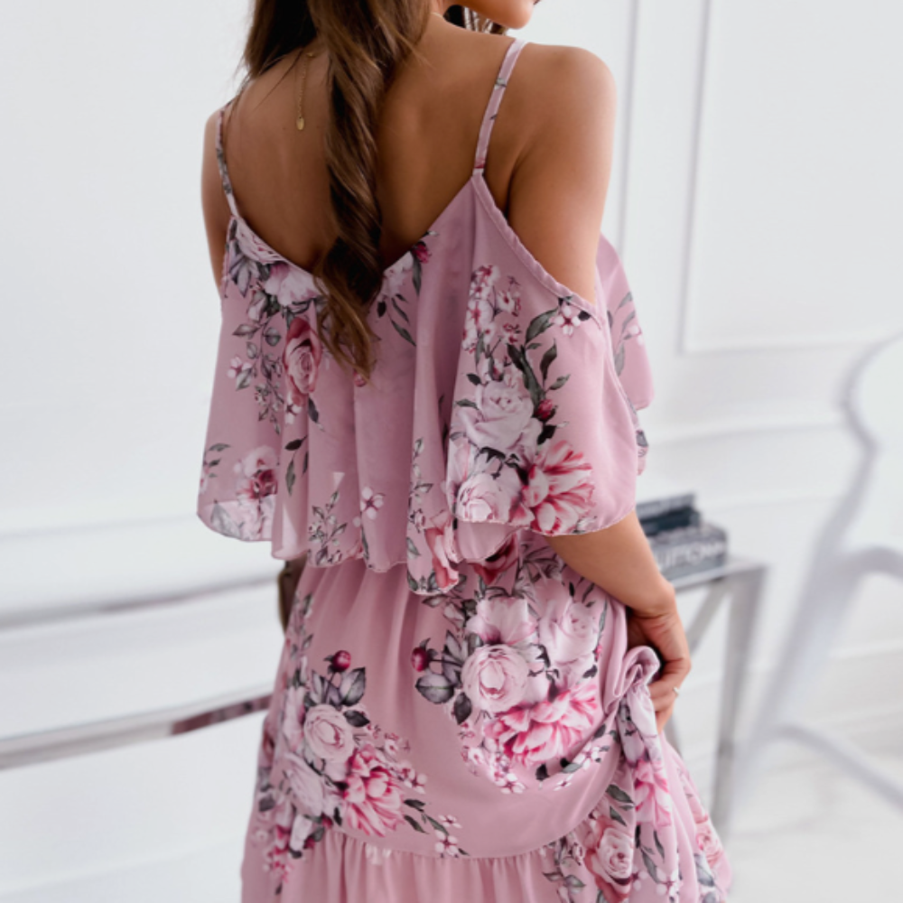 Rear view of a woman wearing a blush pink floral midi dress with off-shoulder ruffle detail and thin straps, showcasing the flowing layered fabric and elegant floral pattern.