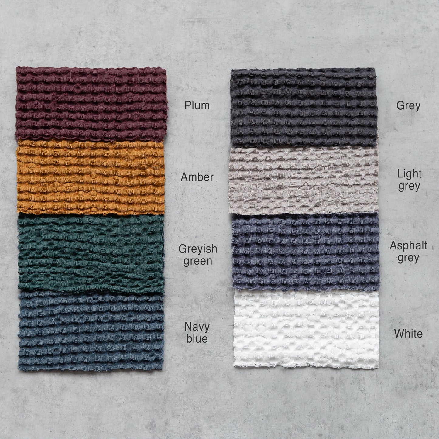 Eight waffle-textured fabric swatches arranged on a concrete surface, labeled with colours: Plum, Amber, Greyish Green, Navy Blue, Grey, Light Grey, Asphalt Grey, and White.