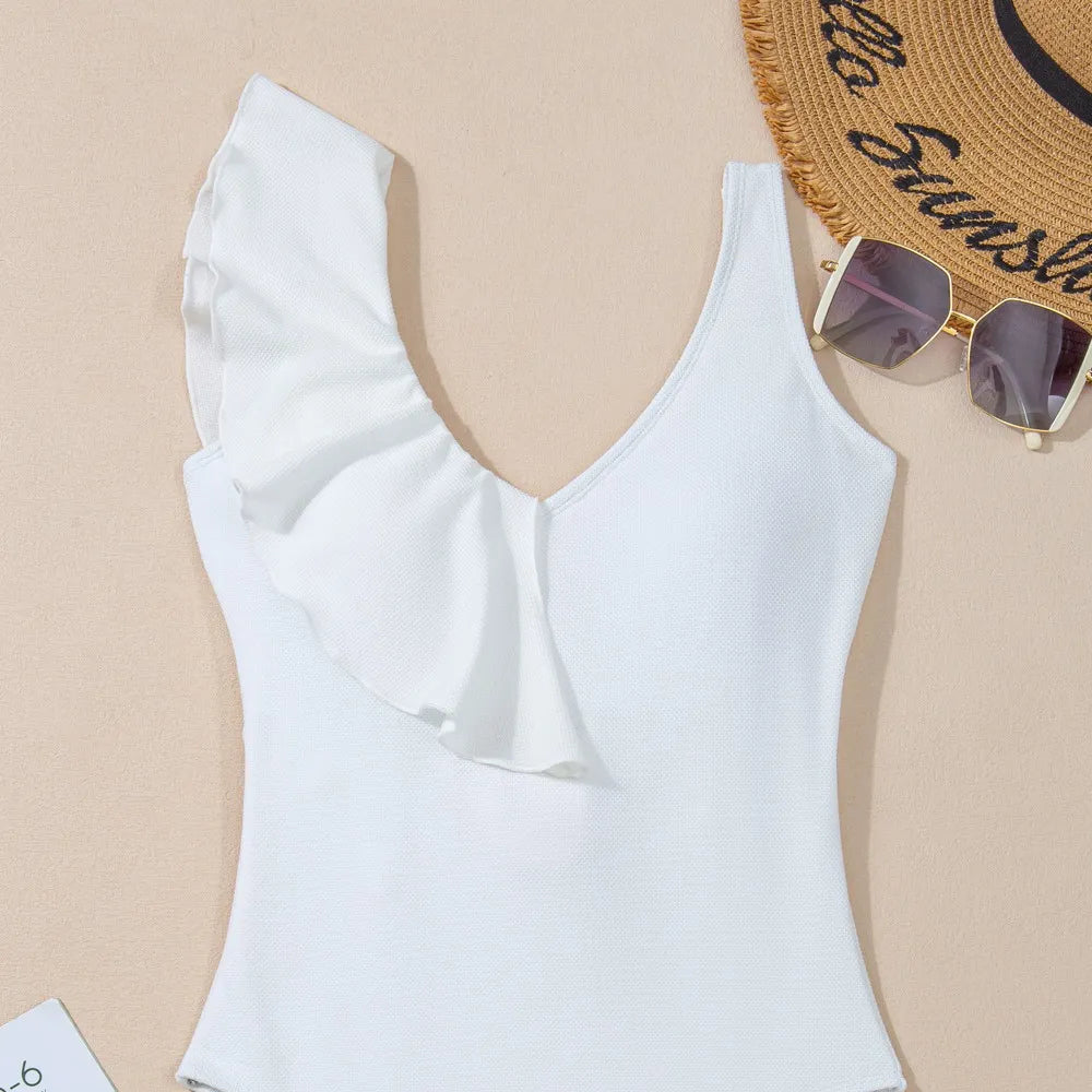 Flat lay of a stylish white one-shoulder ruffle one-piece swimsuit, displayed with sunglasses, a sun hat, and a floral magazine on a beige background.