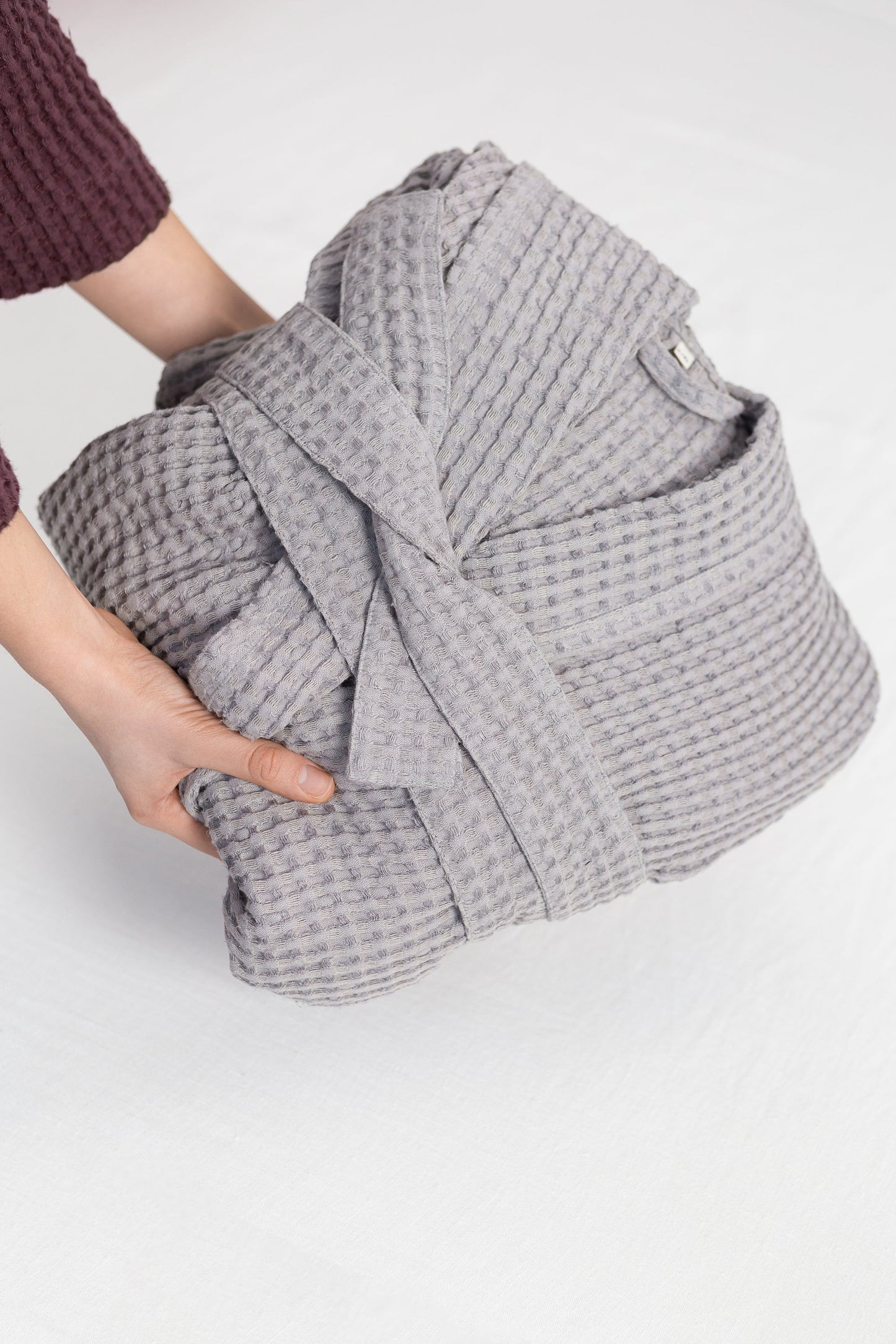Hands holding a neatly folded grey waffle-textured cotton robe with belt tied, showcasing its soft fabric and minimalist design.