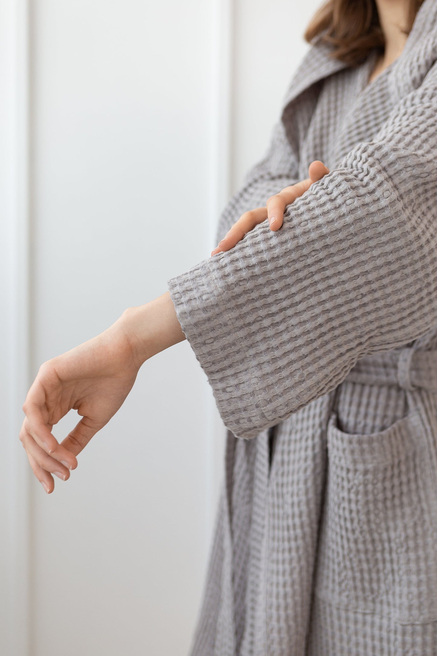 Close-up of woman adjusting the sleeve of a grey waffle-textured cotton bathrobe with tie waist and front pocket.