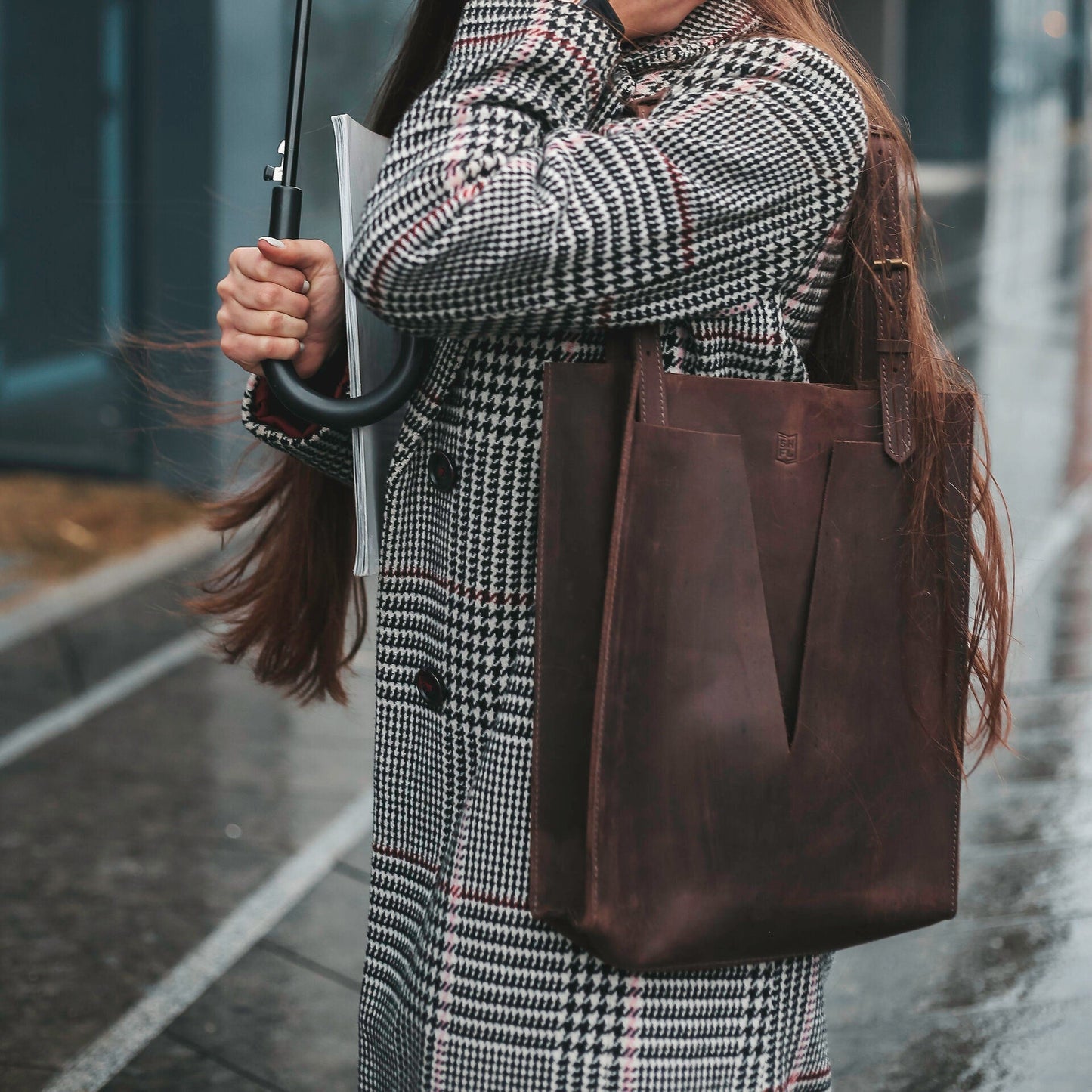 Close-up of a dark brown leather tote bag with minimalist V-cut design, carried over the shoulder of a person in a houndstooth coat, standing on a rainy city street.