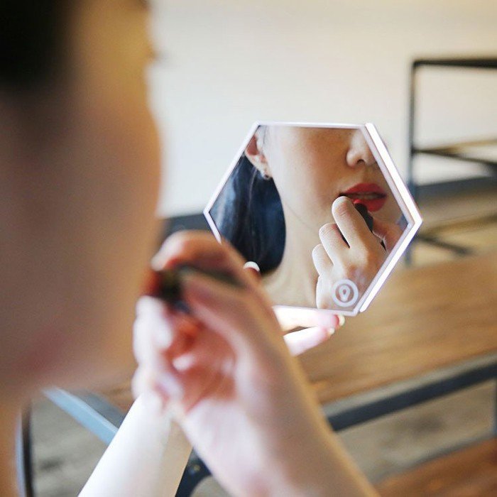 Woman using a hexagonal LED mirror clock to apply lipstick, showcasing the mirror’s smart touch interface and multifunctional design — perfect for beauty routines and modern desk setups.