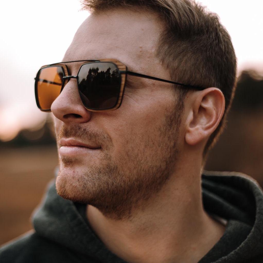 Stylish man wearing Waidzeit wooden-frame sunglasses with amber lenses, reflecting a natural outdoor landscape at sunset.