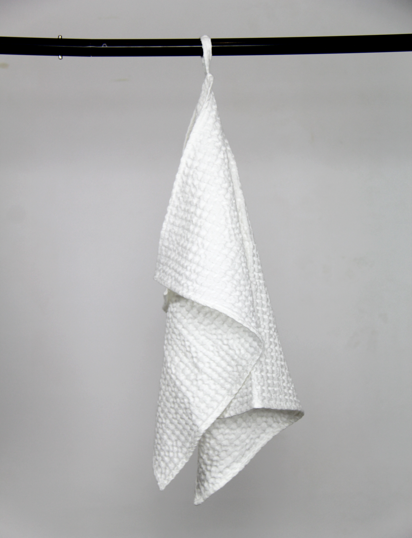 White waffle-textured towel hanging from a black rod by a sewn-in loop, against a plain light grey background