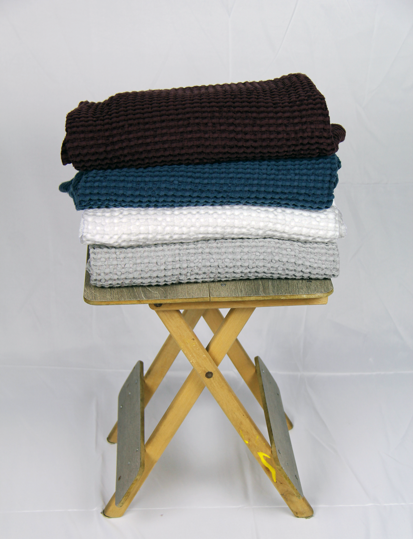 Stack of four folded waffle-textured towels in brown, teal, white, and grey, neatly placed on a rustic wooden folding stool against a plain white background