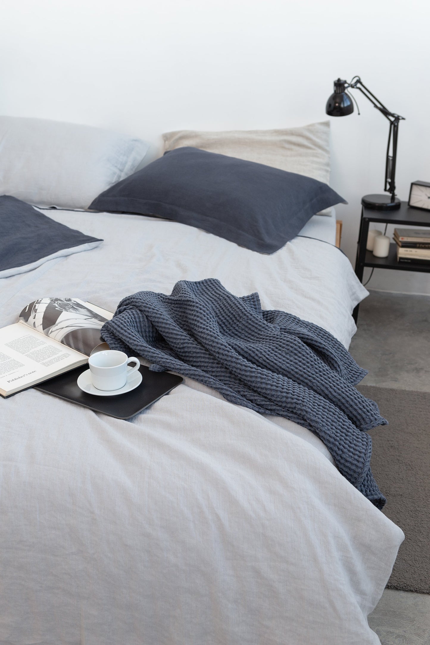 Modern bedroom with light grey bedding, navy blue pillows, a textured dark blue throw blanket, and a black tray holding a book and coffee cup