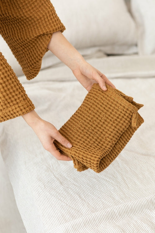 Hands holding folded amber waffle cotton towels on a neutral bedspread — quick-dry, absorbent towels perfect for home, spa, or guest bathrooms
