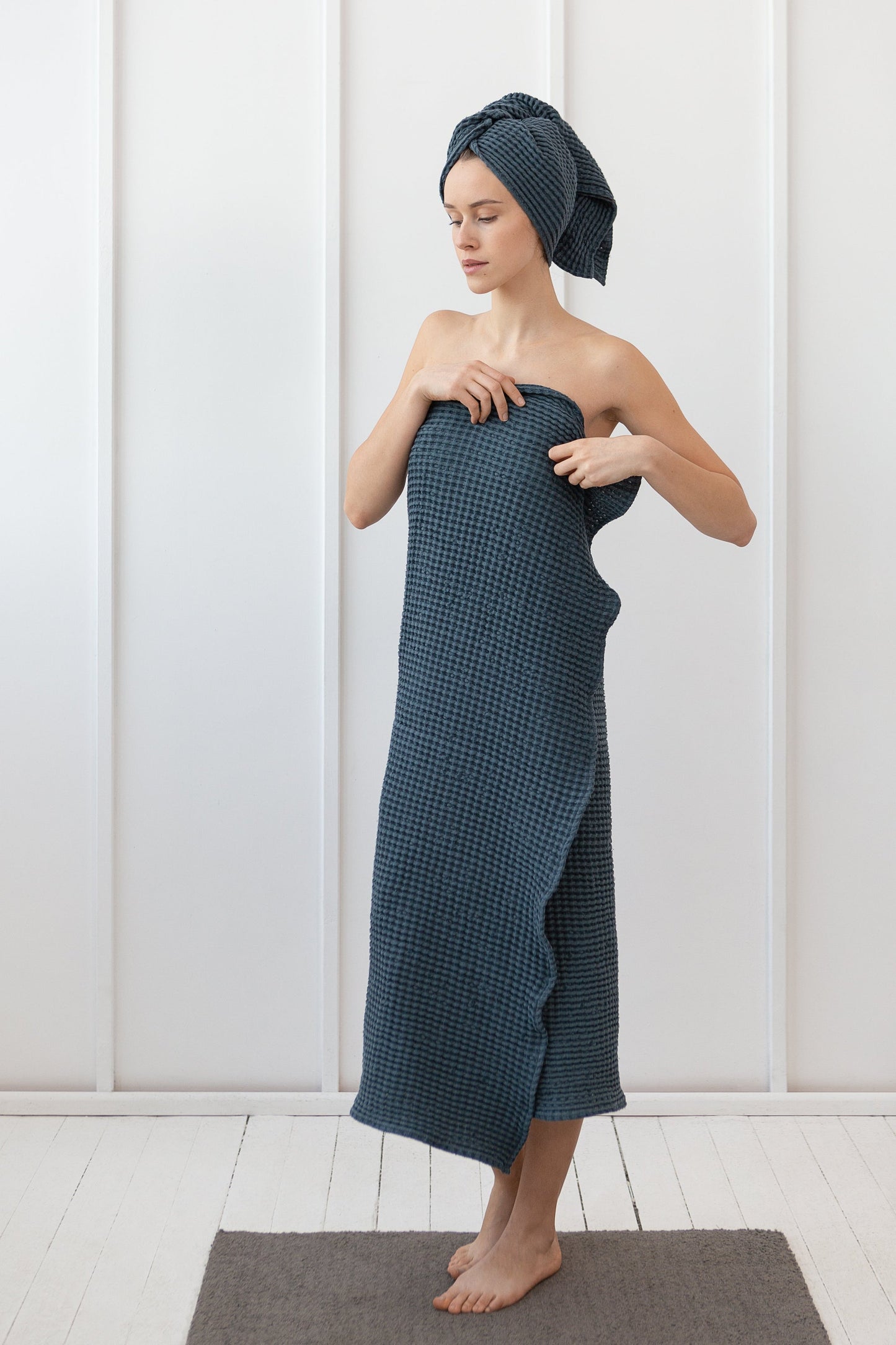 Woman wrapped in a dark blue waffle-textured towel with matching hair wrap, standing on a grey rug in a minimal white room