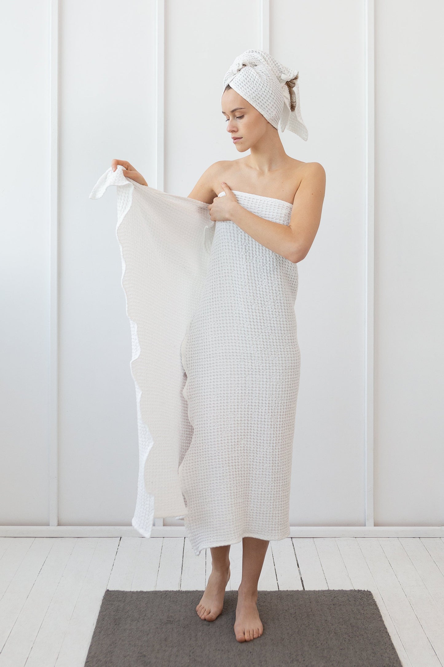 Woman in a white waffle-textured towel wrap and matching hair towel, standing in a minimal white room and holding a second towel