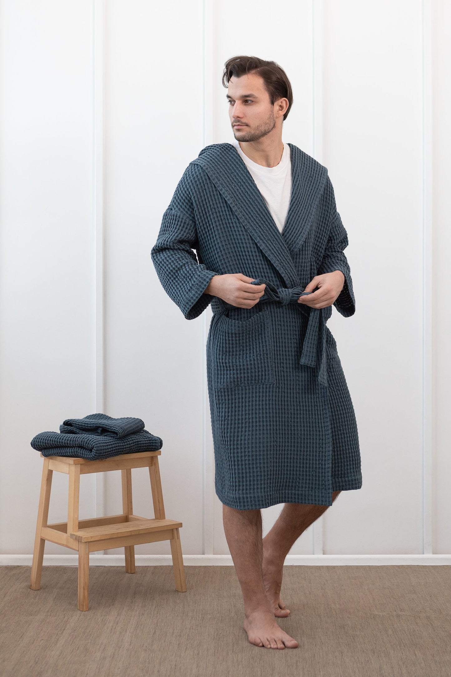 Man wearing a navy waffle cotton bathrobe, tying the belt, standing beside a wooden stool with folded matching robes.