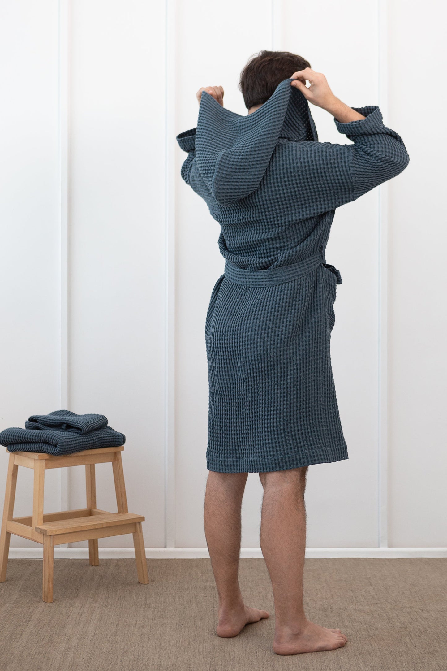 Man wearing a slate blue waffle cotton bathrobe seen from the back, lifting the hood, next to a wooden stool with folded robes.