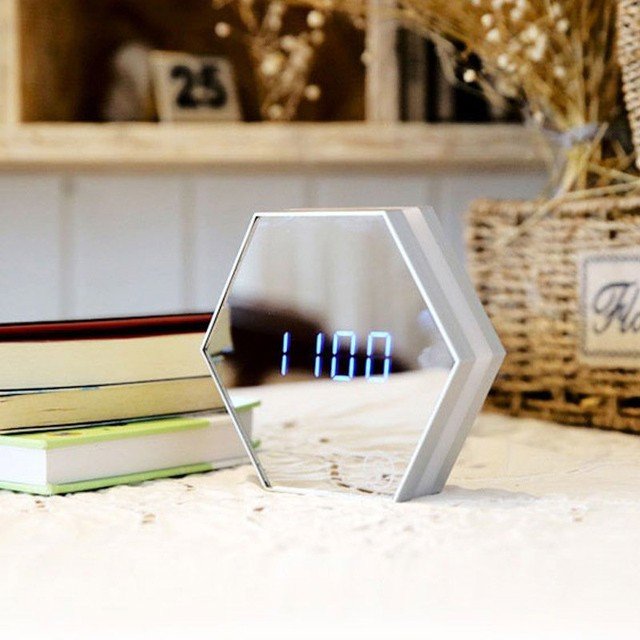 Sleek hexagonal LED mirror clock in white with digital blue time display, placed on a light-toned desk alongside books and rustic décor — ideal for modern home or office environments.