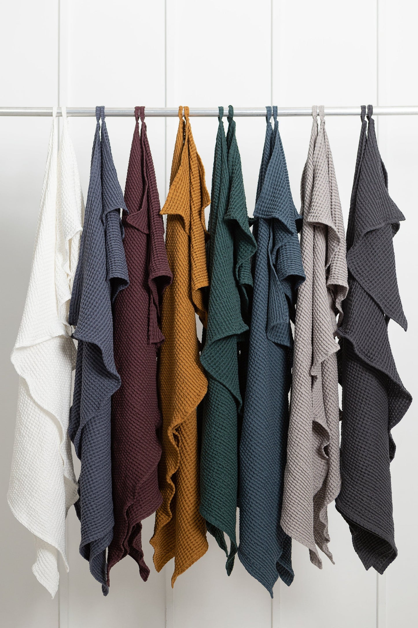 Nine waffle-textured cotton robes hanging on a rail in assorted colours including white, navy, burgundy, mustard, forest green, teal, taupe, slate blue, and charcoal.