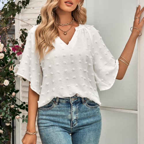 Woman wearing a white Swiss dot V-neck blouse with short flutter sleeves, paired with high-waisted blue jeans and layered necklaces, posing near a floral background.