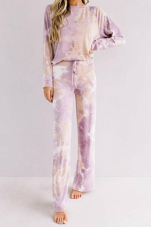 Front view of a woman wearing a cozy two-piece loungewear set featuring a soft lavender and beige tie-dye pattern. The outfit includes a relaxed long-sleeve top and wide-leg drawstring pants. She is barefoot and posed casually against a plain light-colored background.
