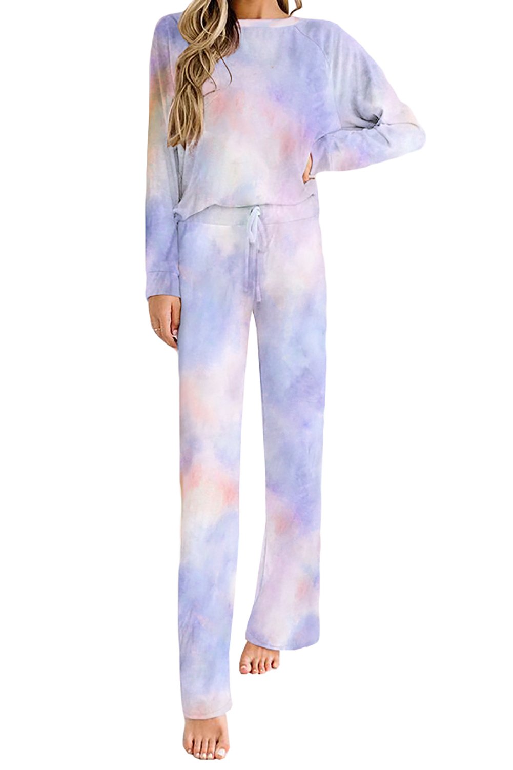 Woman wearing a pastel tie-dye lounge set in shades of blue, purple, and pink, featuring a long-sleeve top and matching drawstring pants. She is standing barefoot against a white background.