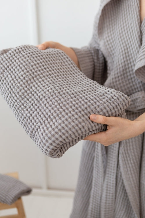 Person holding a folded light grey waffle weave cotton towel while wearing a matching bathrobe — soft and absorbent towel ideal for spa, bath, or home use