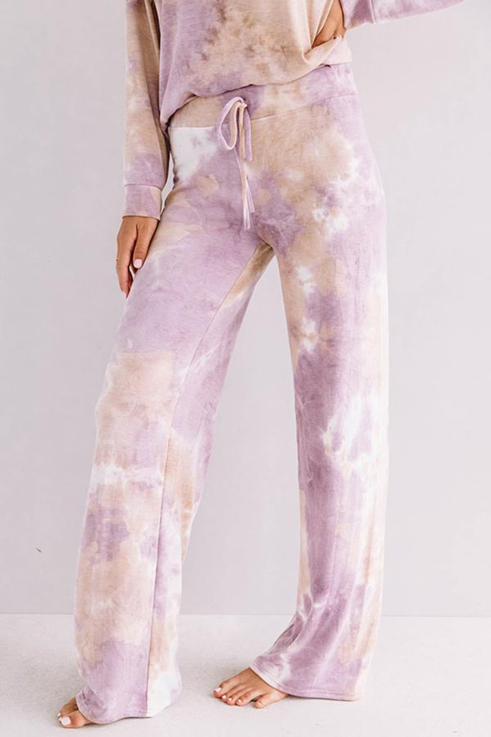 Close-up view of a woman wearing wide-leg lounge pants with a drawstring waist in a soft lavender and beige tie-dye pattern. She is barefoot and standing against a light background, with the matching long-sleeve top partially visible.