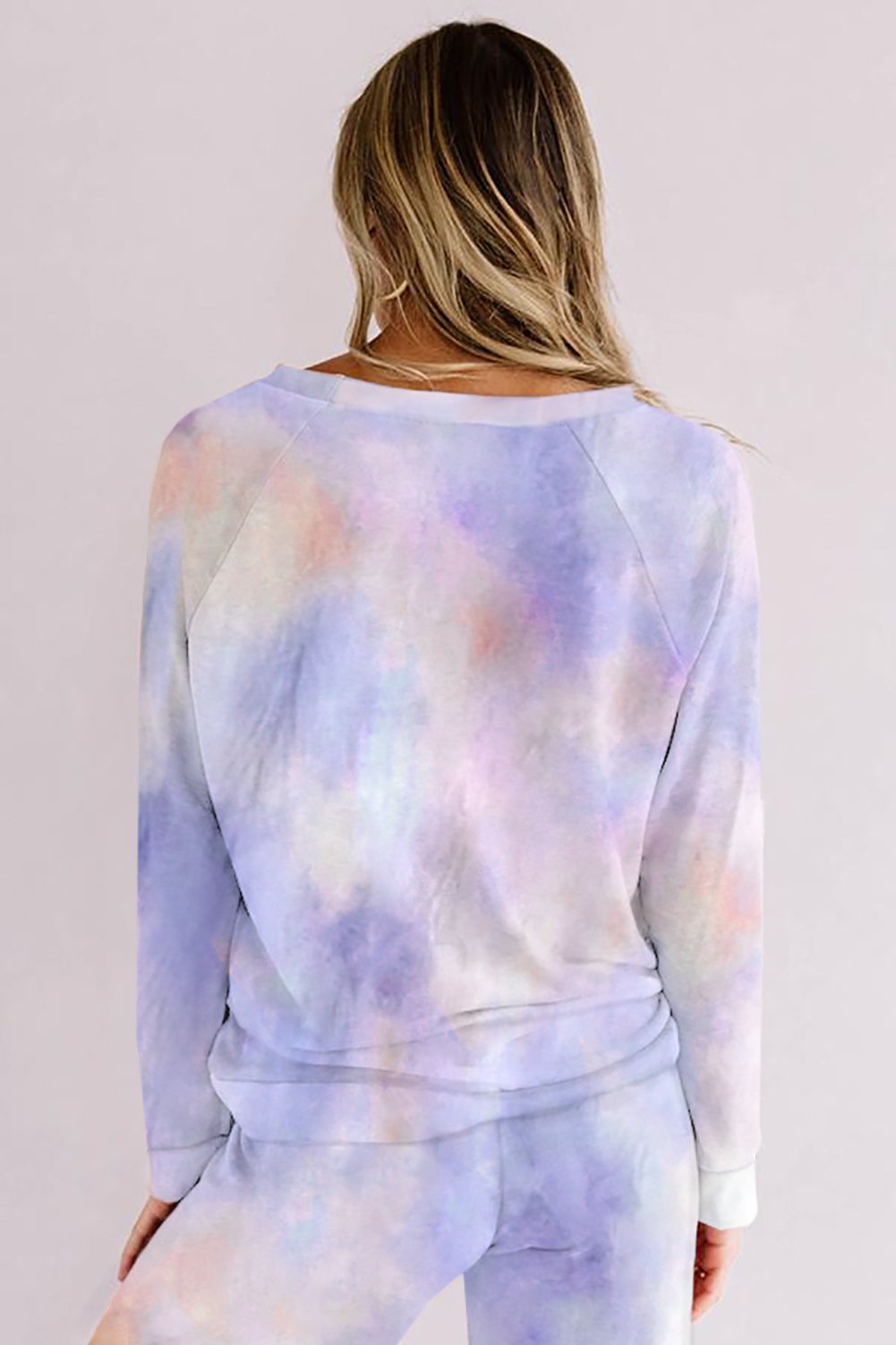 Back view of a woman with long blonde hair wearing a pastel tie-dye loungewear set in shades of lavender, peach, and blue. She stands against a pale background, showcasing the relaxed fit and soft texture of the outfit.