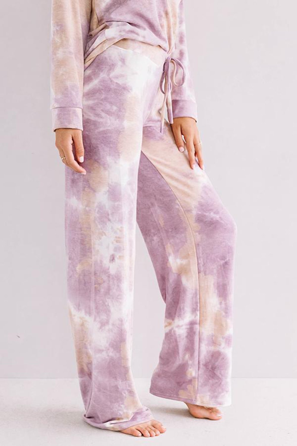 Close-up of a woman's legs wearing mauve and beige tie-dye loungewear pants with a drawstring waist. The pants are full-length with a relaxed fit, and she is barefoot, standing against a light background.