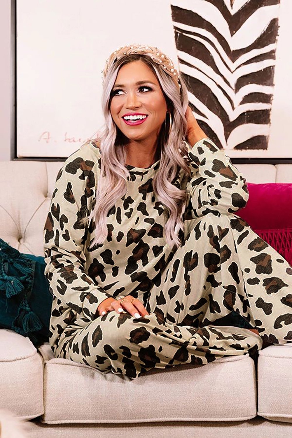Smiling woman with long platinum blonde waves sits cross-legged on a cream sofa, wearing a matching leopard-print lounge set in neutral tones. She accessorizes with a pearl headband and is surrounded by plush cushions and a zebra-print wall art.