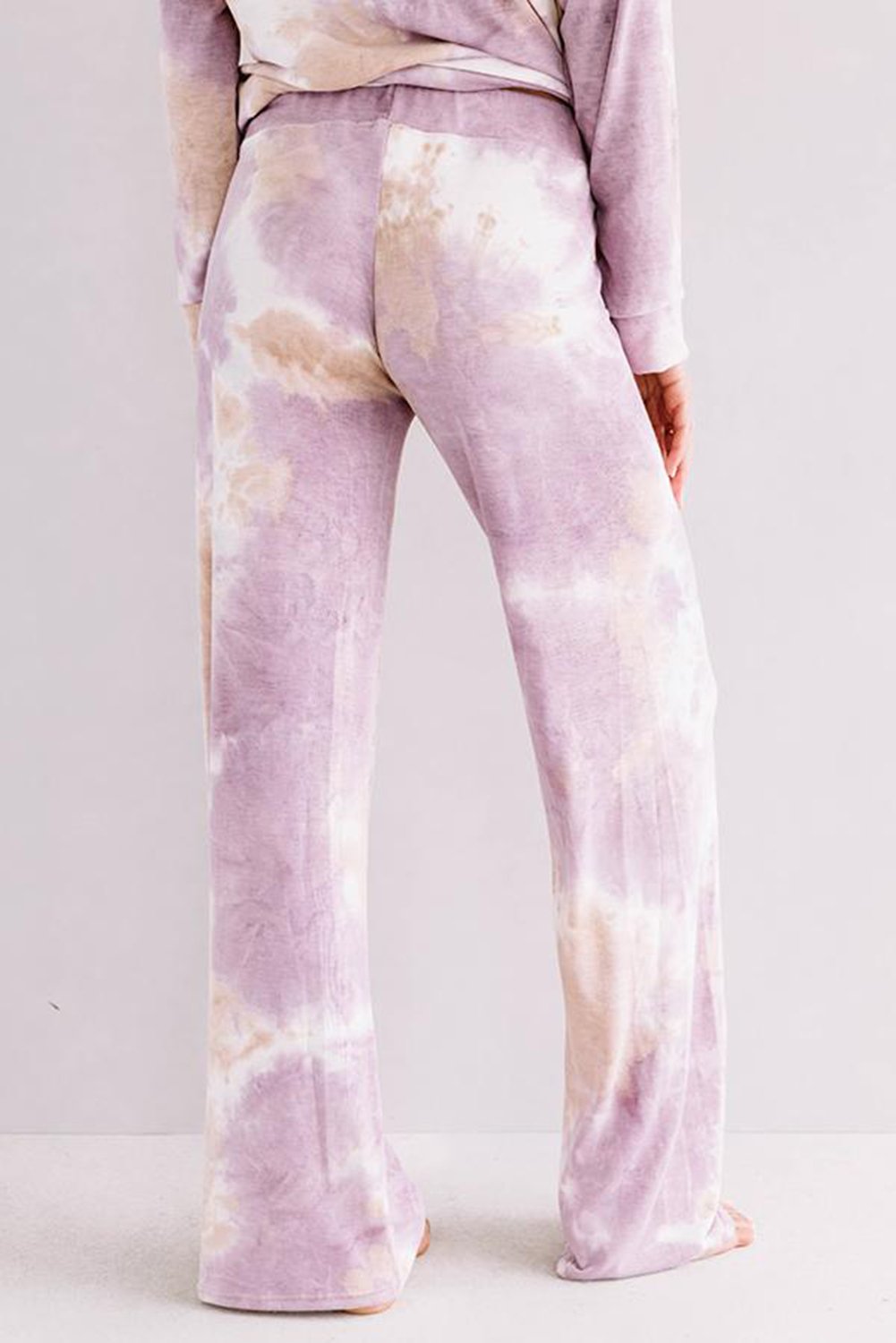 Rear view of a person wearing soft, wide-leg lounge pants in a pastel purple and beige tie-dye print. The fabric appears plush and cozy, and the pants are paired with a matching long-sleeve top. The person is barefoot and standing against a light-colored background.