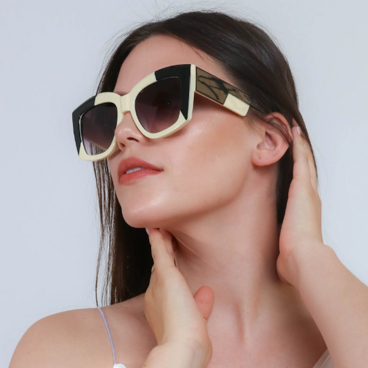 Woman wearing oversized square sunglasses with a bold black and ivory frame, posing with a hand on her neck and long dark hair tucked behind her ear.