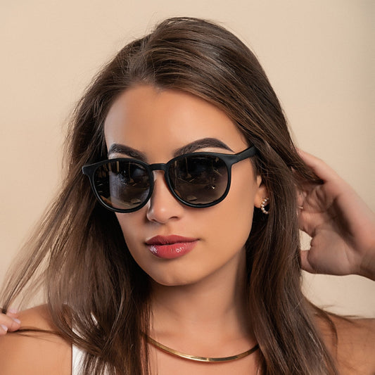 Woman wearing classic black sunglasses and a white tank top, posing confidently with one hand in her hair and a gold necklace around her neck.