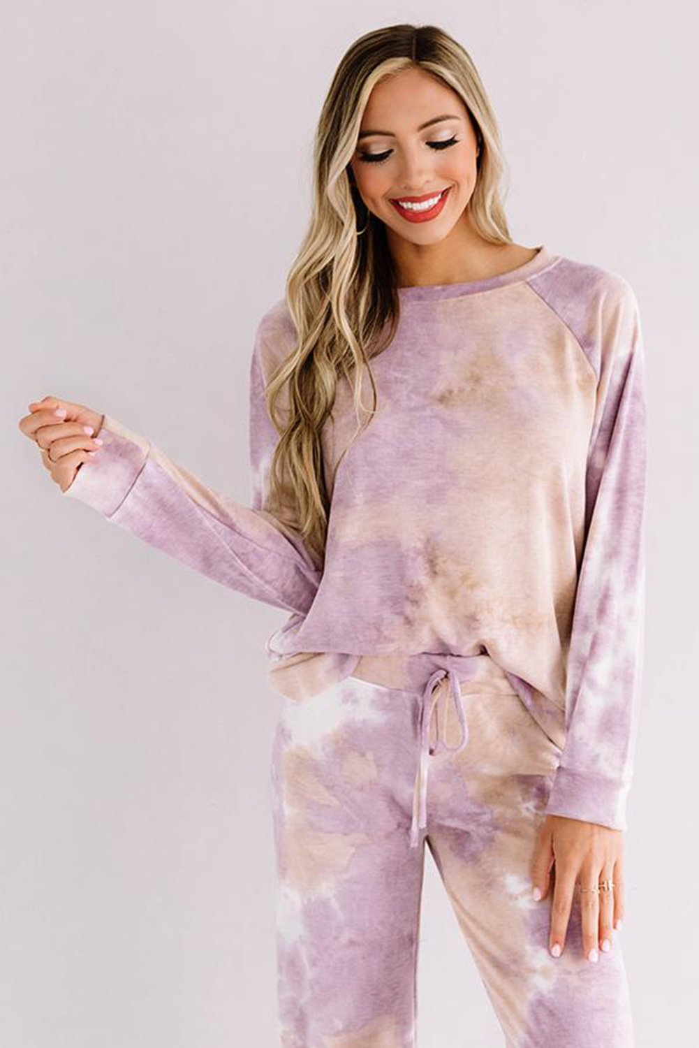 Woman wearing a soft mauve and beige tie-dye loungewear set, featuring a relaxed long-sleeve top and drawstring pants. She is smiling with one hand raised, giving a casual, cozy vibe.