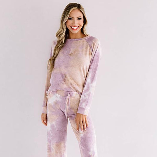 Smiling woman standing barefoot against a white background, wearing a soft pastel tie-dye lounge set with long-sleeve top and wide-leg drawstring pants in shades of lilac, beige, and ivory