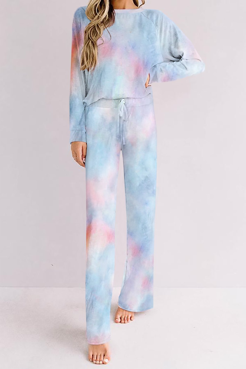 Woman standing barefoot against a pale background, wearing a soft pastel tie-dye lounge set with long-sleeve top and drawstring wide-leg pants in a dreamy blend of light blue, pink, purple, and hints of peach.