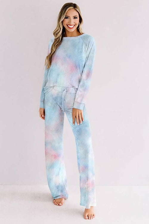 Smiling woman in a pastel blue and pink tie-dye loungewear set, featuring a long-sleeve top and matching drawstring wide-leg pants. She is barefoot and standing in a bright, minimalist indoor space with a soft, neutral background.