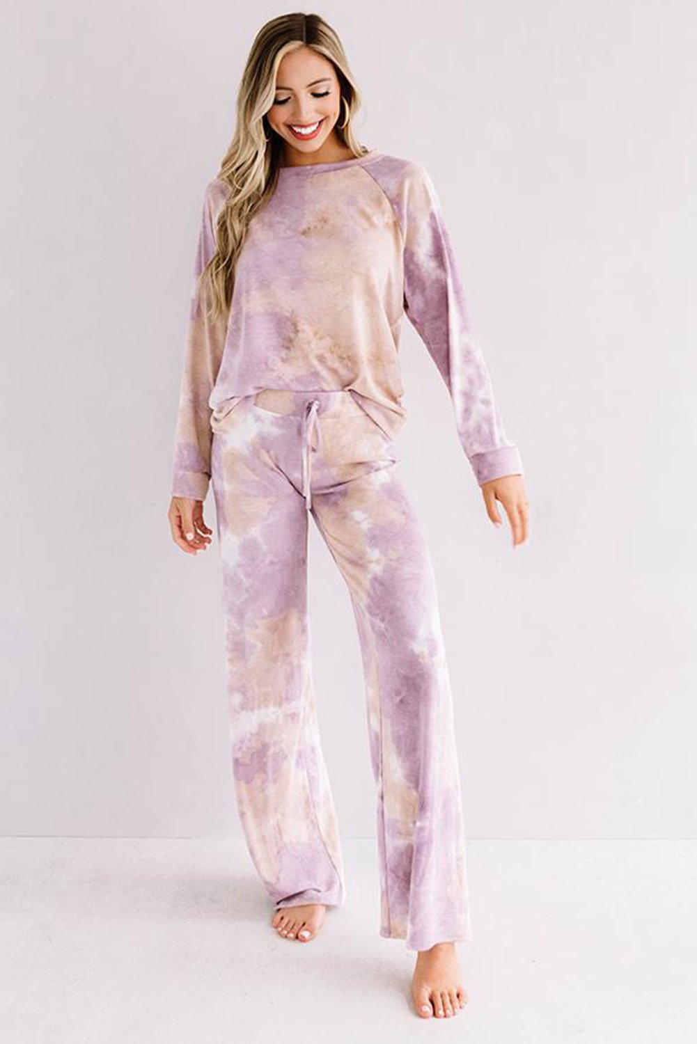 Woman wearing a soft, pastel tie-dye lounge set in shades of lilac and beige. The outfit includes a long-sleeve pullover and drawstring wide-leg pants, styled for a cozy and relaxed look. She stands barefoot against a light pink wall, smiling and looking down.