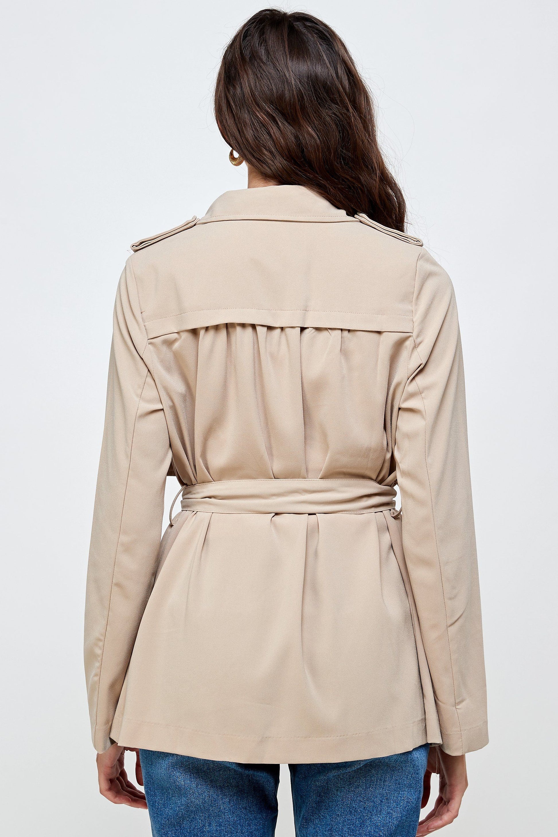 Rear view of a woman wearing a beige belted trench jacket with a storm flap and pleated waist, paired with denim jeans.
