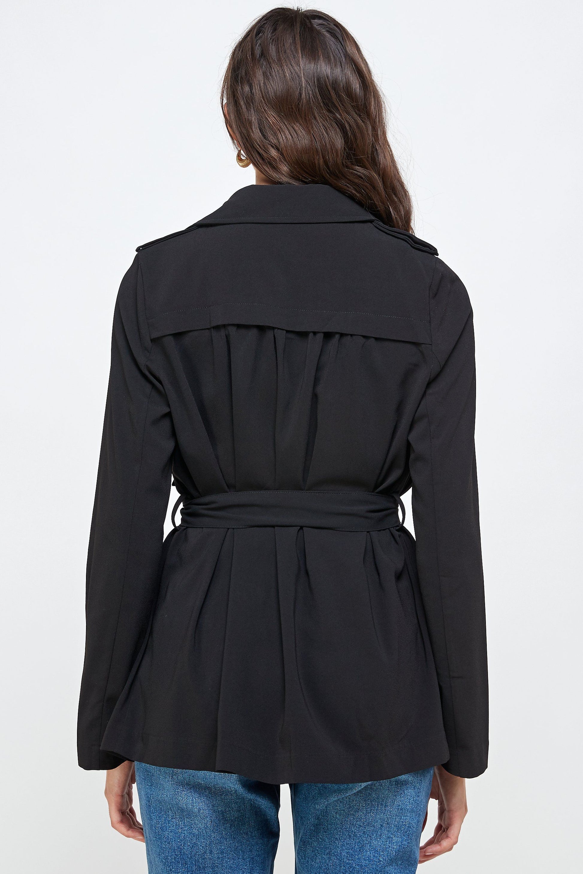 Rear view of a woman wearing a black belted trench jacket with a back storm flap and gathered waist detail, styled with denim jeans.