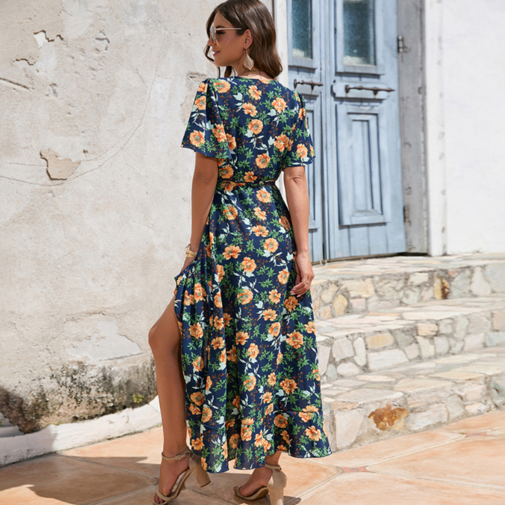 Rear view of woman in a navy floral wrap dress with side slit, wearing heels and sunglasses, standing near a rustic stone staircase in sunlight.