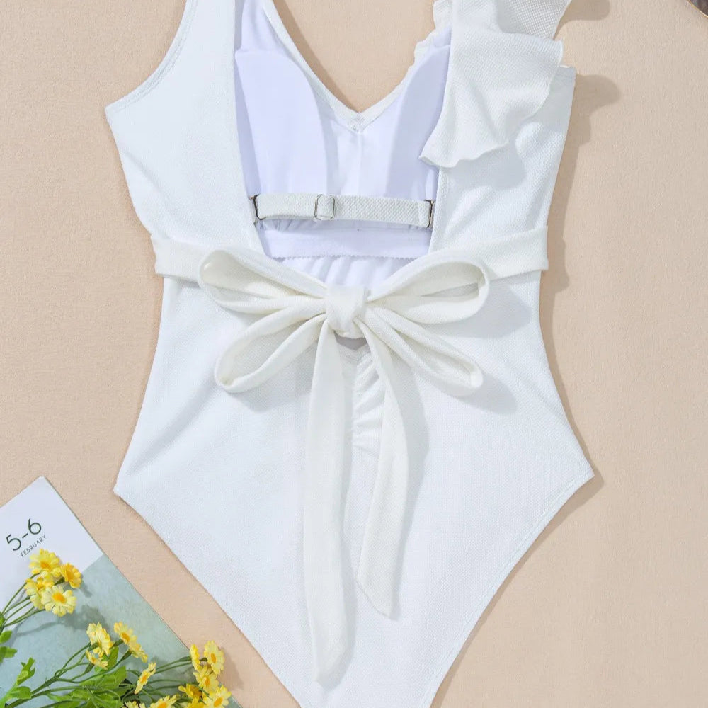 Flat lay showing the back of a white one-shoulder ruffle one-piece swimsuit with waist tie detail and buckle closure, styled with sunglasses, a sun hat, and a floral magazine.