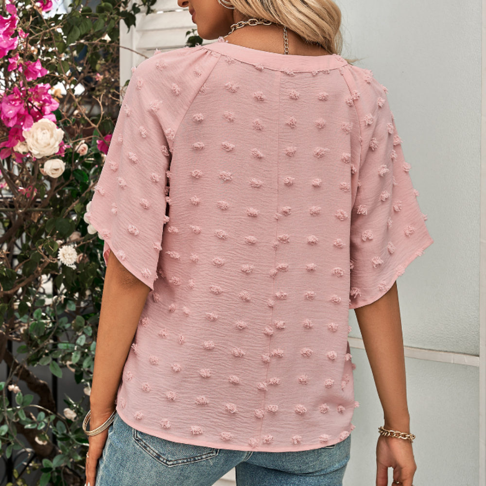 Rear view of woman wearing a light pink Swiss dot textured blouse with short sleeves and relaxed fit, paired with denim jeans and gold accessories.