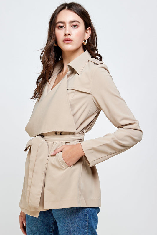 Woman wearing a beige belted trench jacket with wide lapels, flap detailing, and long sleeves, styled with blue denim jeans and gold hoop earrings against a neutral background.