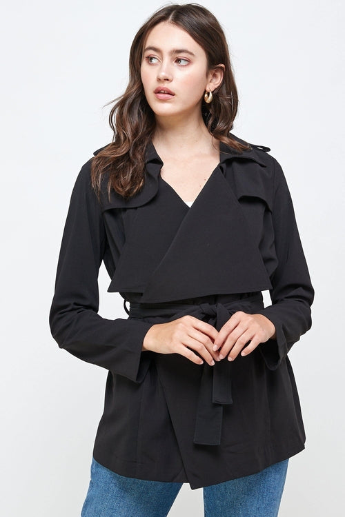 Woman wearing a black belted trench jacket featuring oversized lapels, waist tie, and long sleeves, styled with blue jeans.