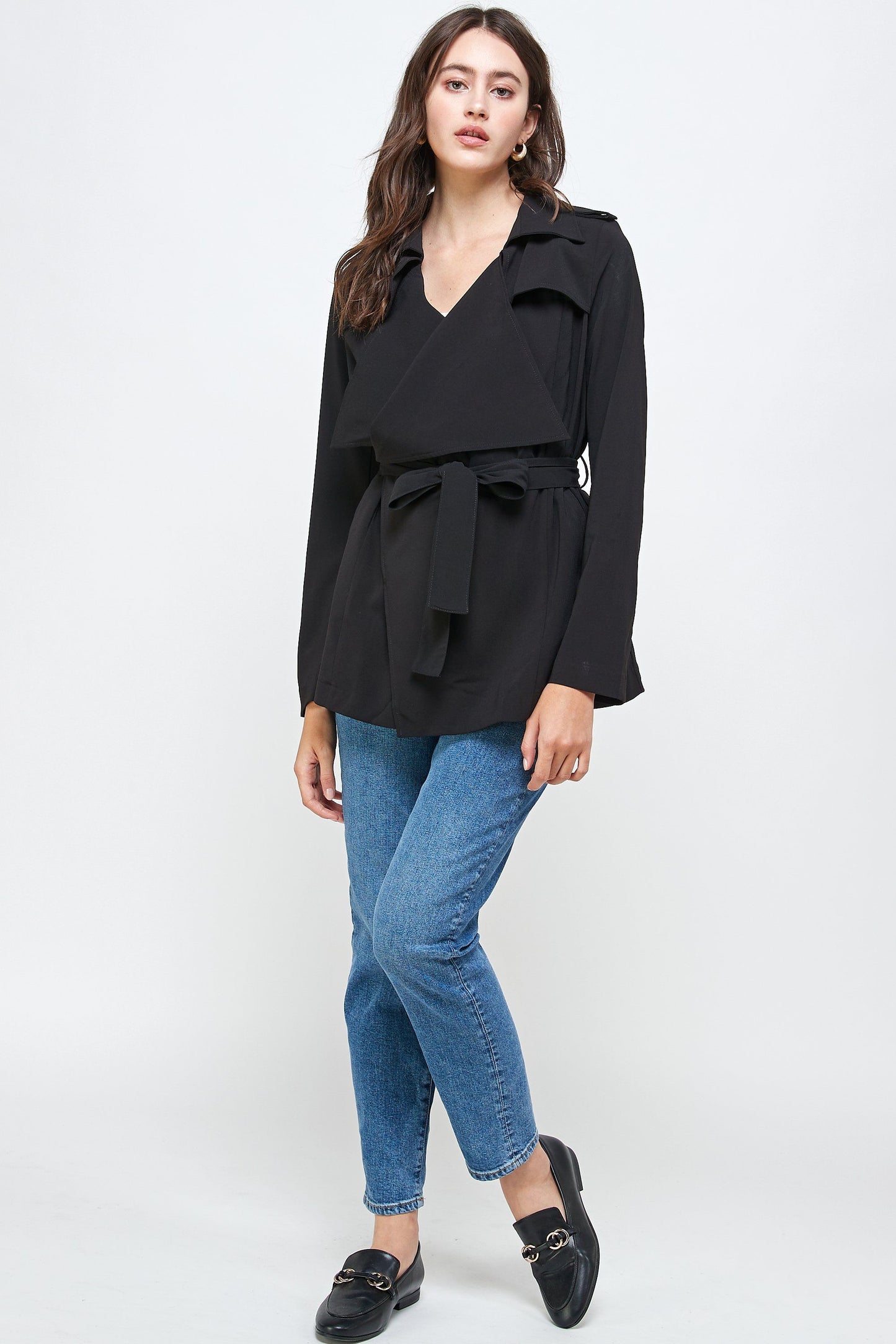 Woman wearing a stylish black belted trench jacket with wide lapels and long sleeves, paired with blue jeans and black loafers, posing against a plain background.