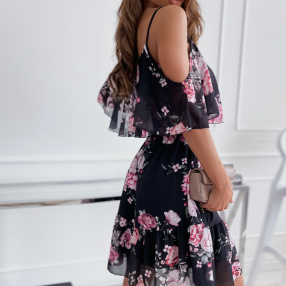 Side view of a woman wearing a black midi dress with pink floral print, cold shoulder cutouts, ruffled overlay, and spaghetti straps – styled with a neutral clutch.